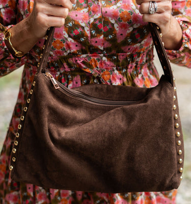 Brown handbag with gold studs held by a person wearing a colorful floral dress.