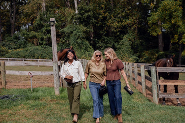 Three women walking together in a grassy area with wooden fences and horses in the background.